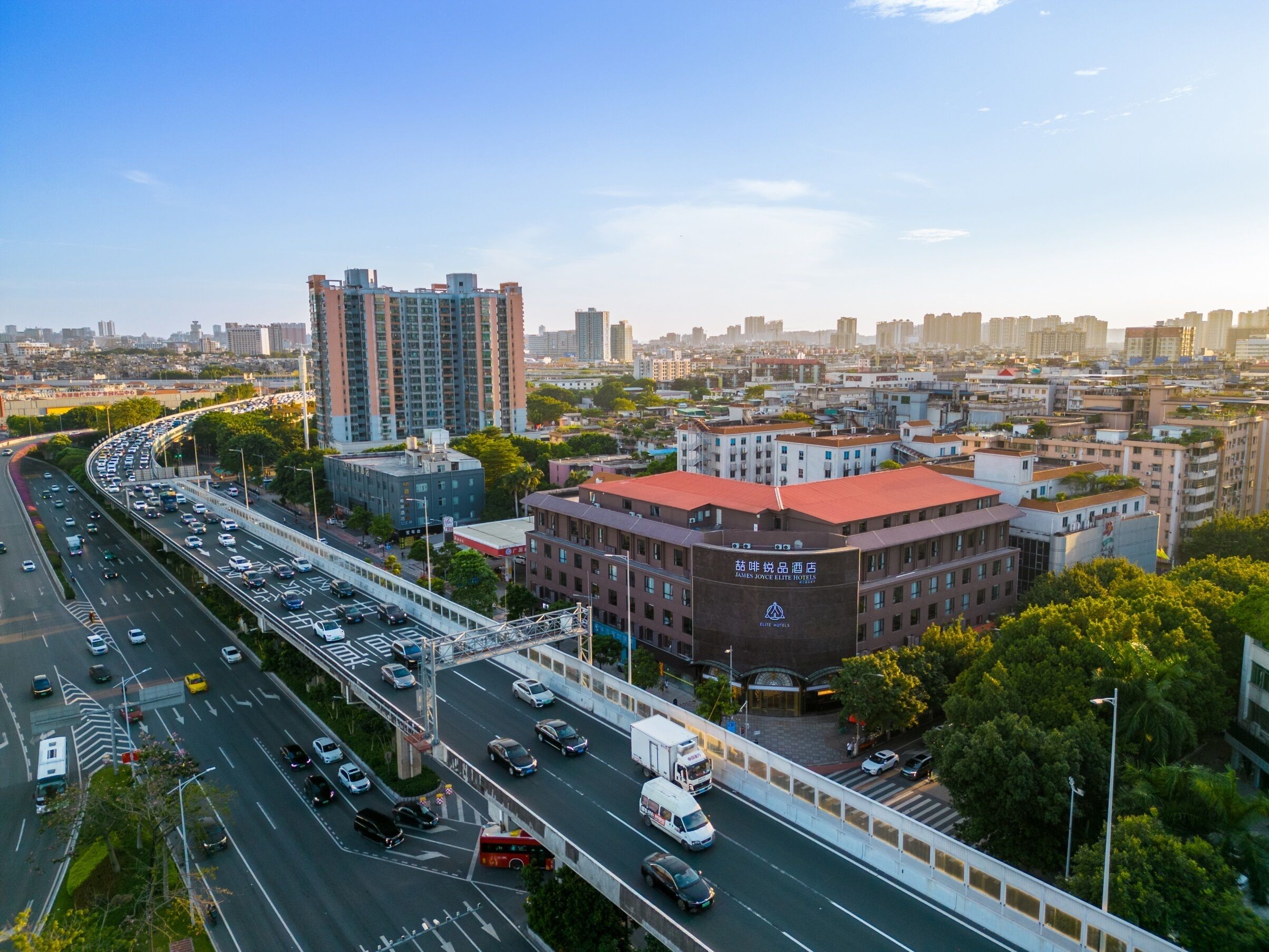 A multi-level hotel with a red roof, surrounded by a cityscape with high-rise buildings and a busy highway.