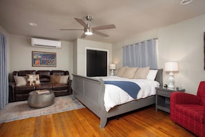 A bedroom with a bed, a sofa, a red chair, a nightstand, a lamp, an air conditioner, and a ceiling fan.