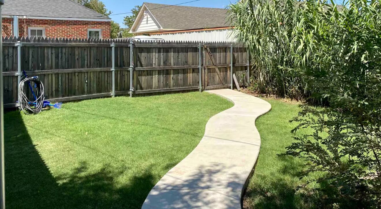A backyard with a curved concrete pathway, a wooden fence, and a grassy lawn.