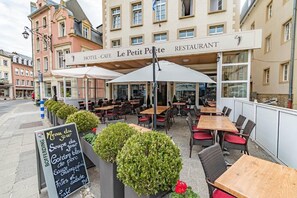 An outdoor cafe with tables and chairs, a menu board, and a building with a sign that reads 'Le Petit Poete Restaurant'.