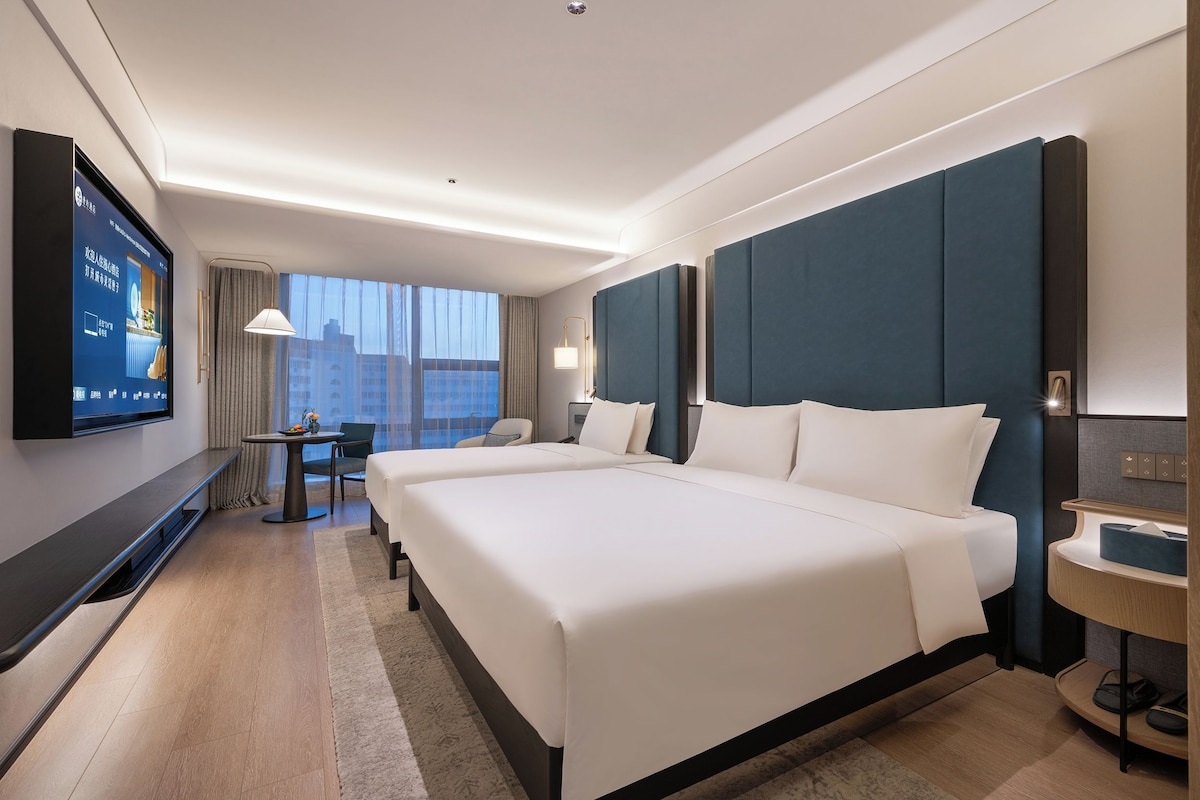 Quarto de hotel com uma cama grande, TV na parede, escrivaninha e janela com vista para a cidade.
