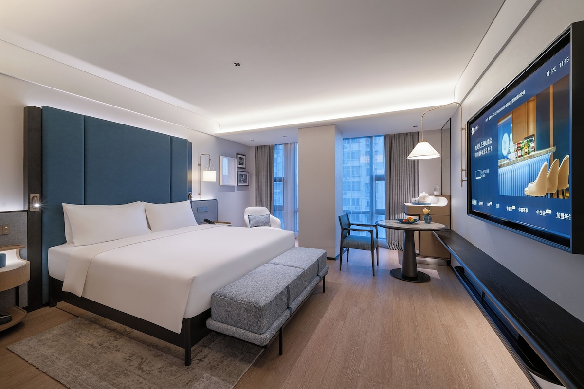 Quarto de hotel com uma cama grande, uma escrivaninha, uma cadeira, uma televisão e uma janela.