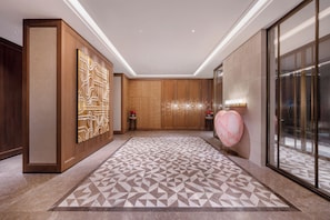 Un moderno vestíbulo de hotel con alfombra de estampado geométrico, paneles de madera y un gran ascensor de vidrio.