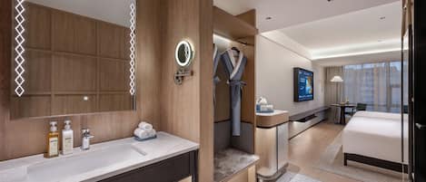 Habitación de hotel moderna con un espejo grande, tocador con artículos de aseo, zona de ducha, cama, televisor y vistas a la ciudad.