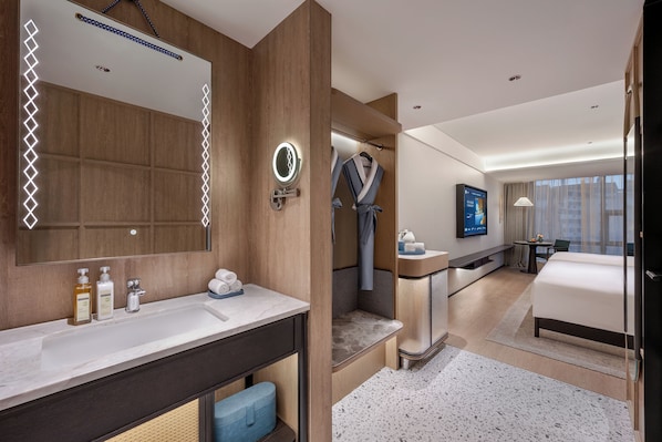 Habitación de hotel moderna con un espejo grande, tocador con artículos de aseo, zona de ducha, cama, televisor y vistas a la ciudad.