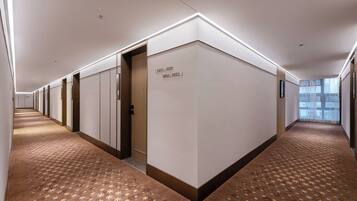 Corredor de hotel com carpete estampado, porta numerada e janela com vista.