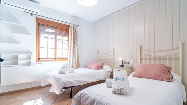 Una camera d'albergo con due letti, una finestra con tende, un comodino con lampada e una mensola a muro.