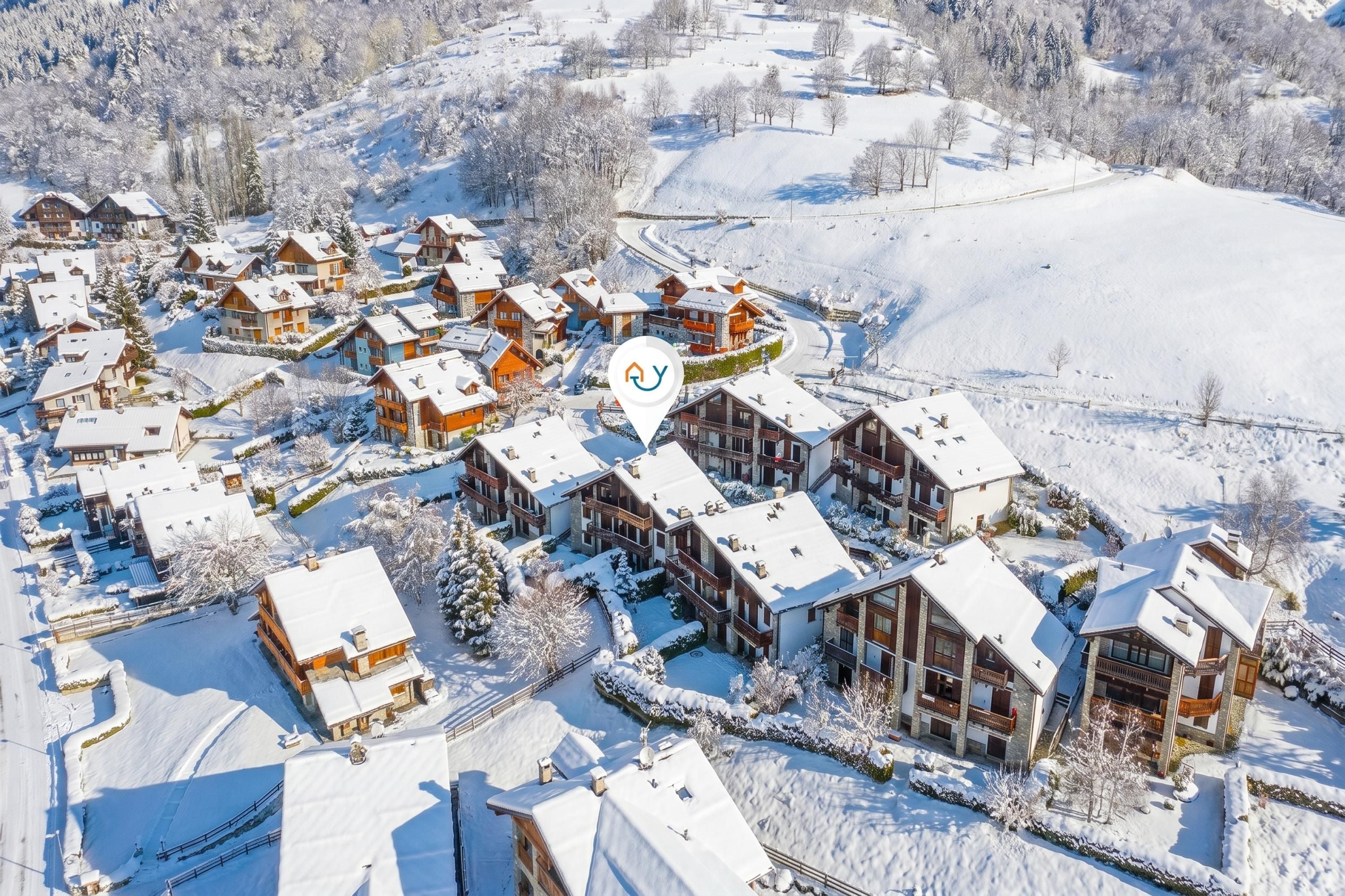 A snow-covered village with multiple chalet-style buildings, a winding road, and a prominent ski lift.