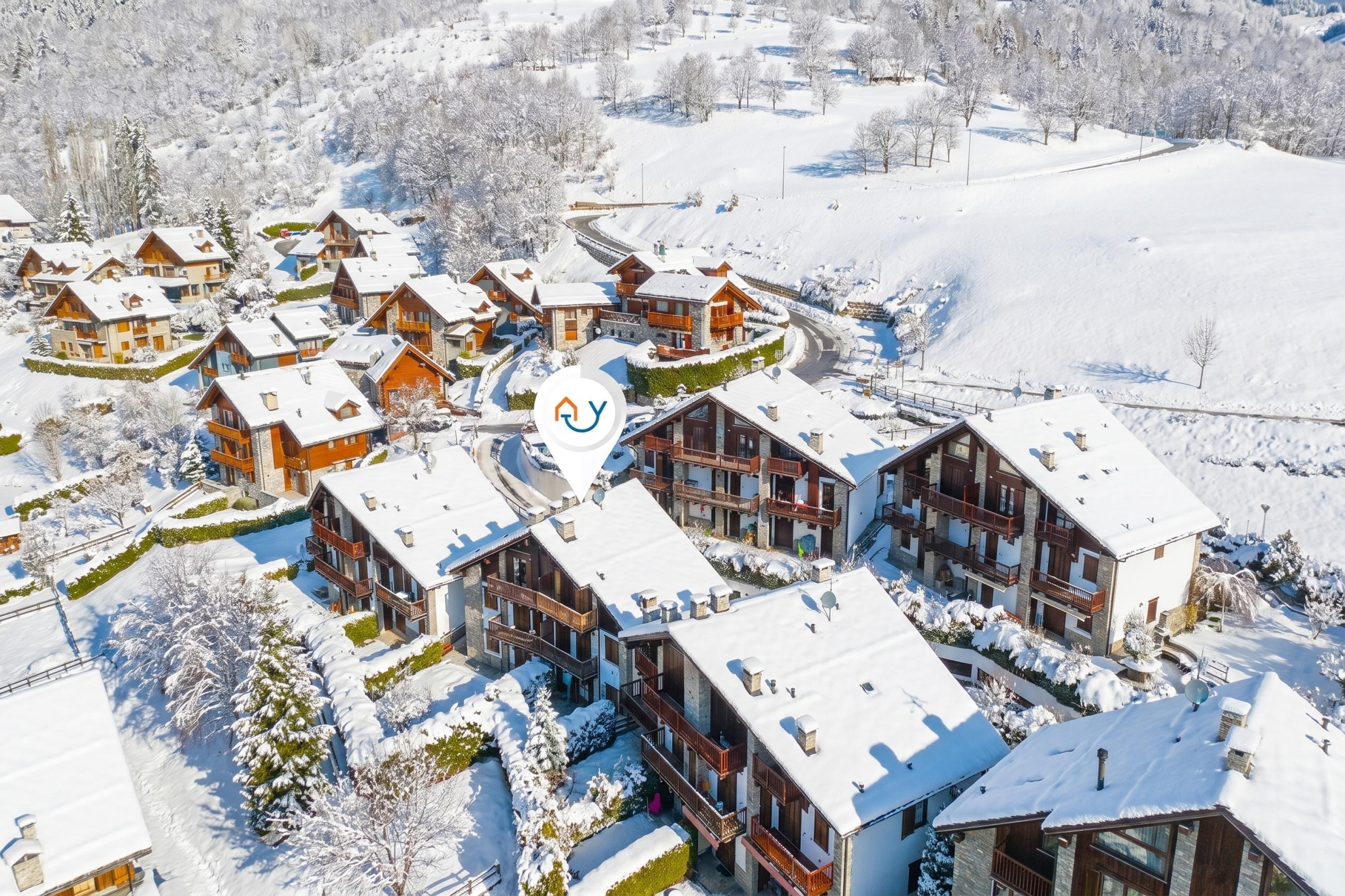 A snow-covered resort with multiple chalet-style buildings, a central communal area, and a ski slope in the background.