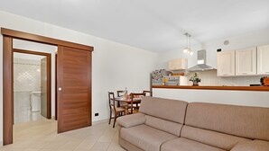 A modern kitchen with white cabinets, a dining area with a wooden table and chairs, and a beige sofa in a living room. - Appartamento Fior di Roccia (Bardonecchia)