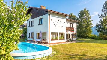 A two-story house with a swimming pool, a patio with outdoor furniture, and a large clock on the front wall.