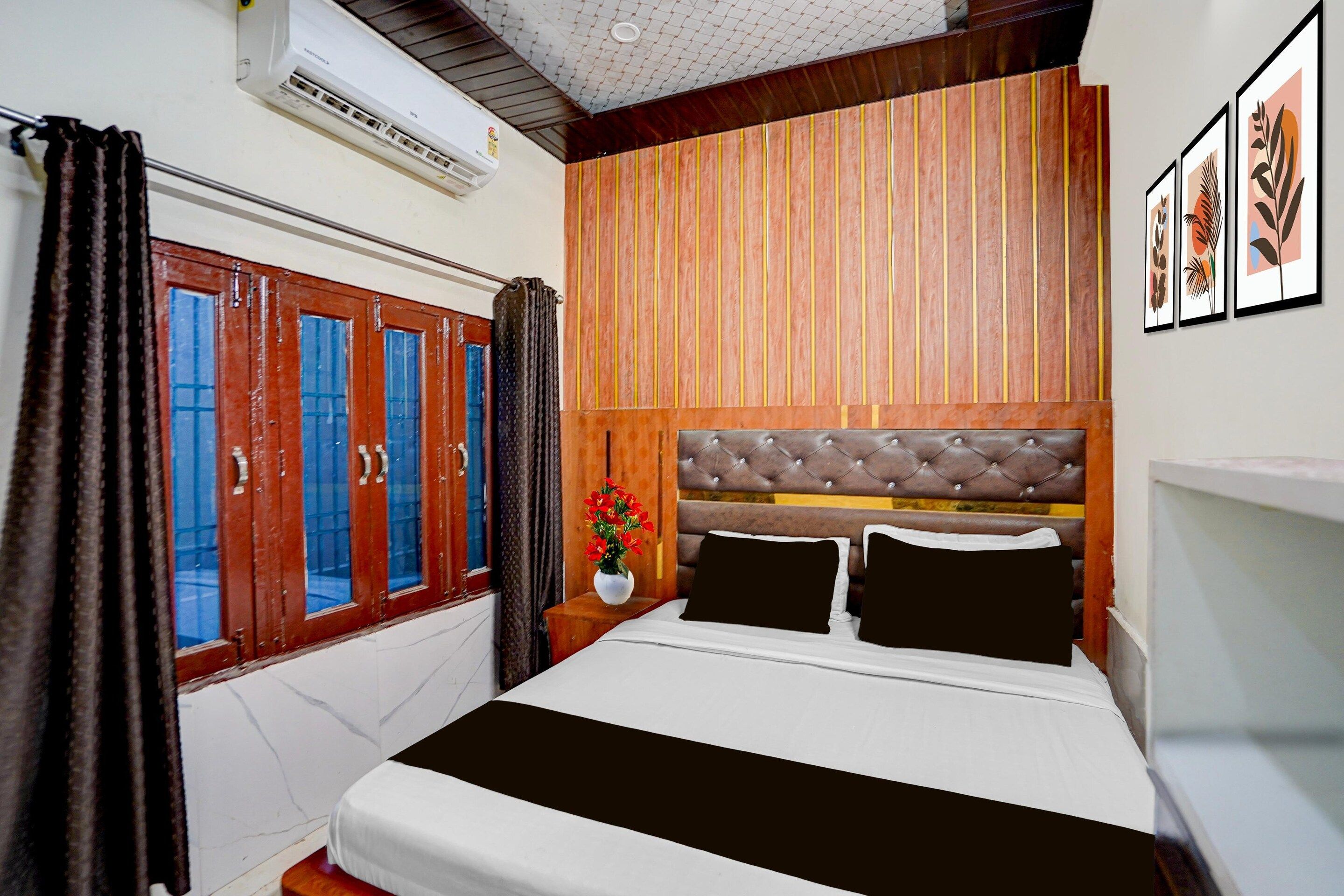 Une chambre d’hôtel comprenant un lit, des rideaux, une fenêtre, un appareil de climatisation, une étagère fixée au mur et des œuvres d’art encadrées.