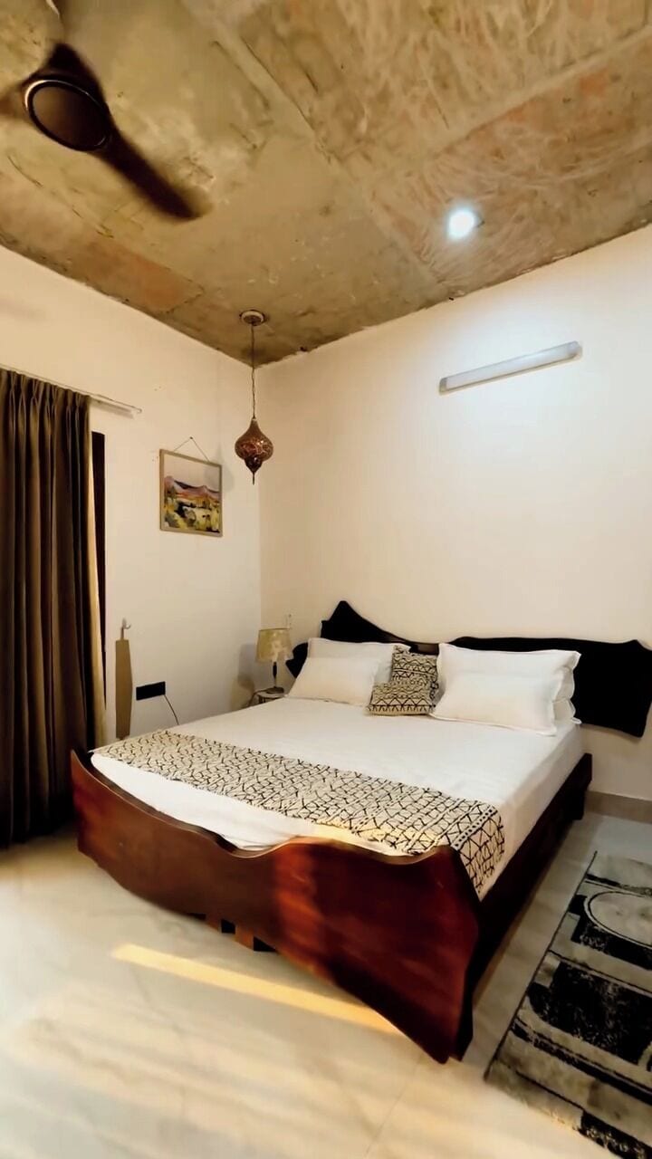 Un dormitorio con cama, ventilador de techo, lámpara colgante, un cuadro y una alfombra.
