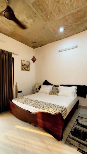 A bedroom with a bed, a ceiling fan, a hanging light fixture, a painting, and a rug. - Abhyaranyan Farmstay (Behat)