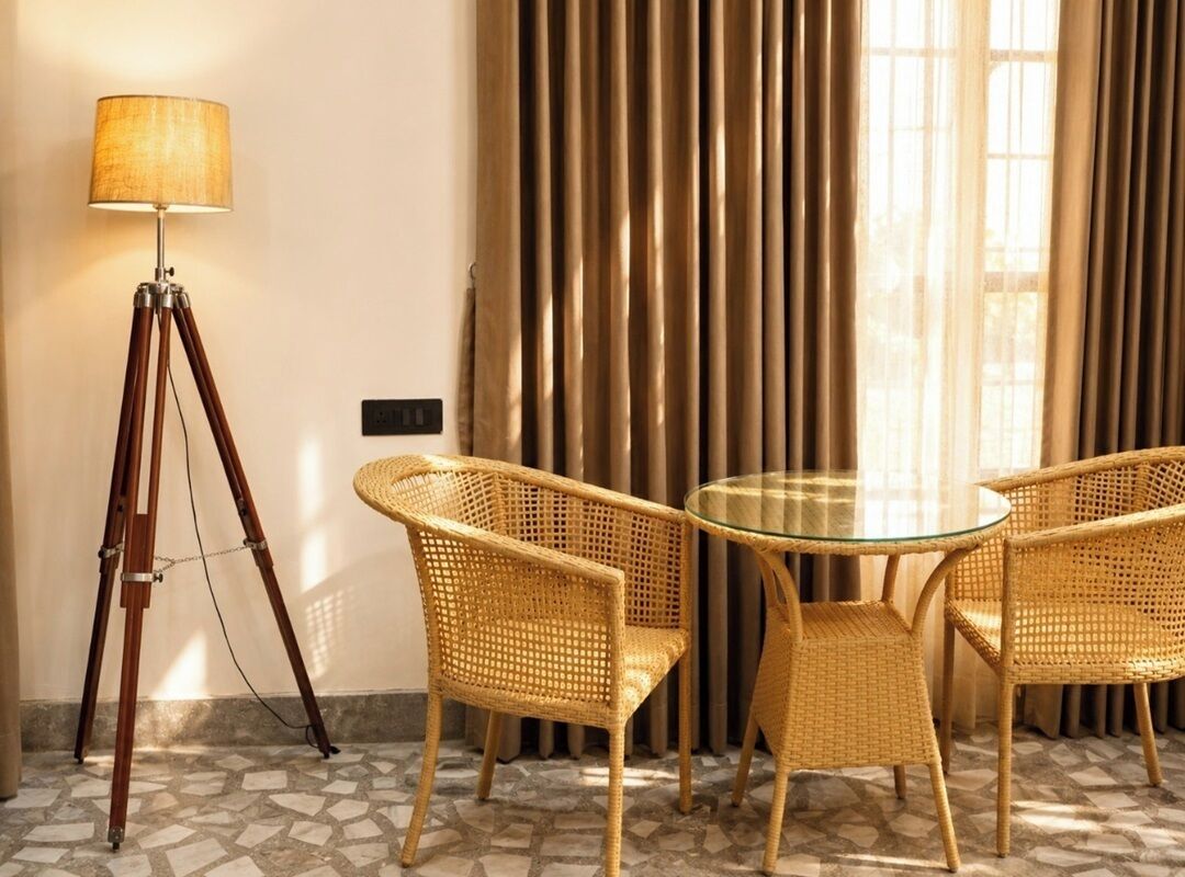 Una habitación con lámpara de pie con trípode, sillas de mimbre, una mesa de vidrio y una ventana con cortinas.