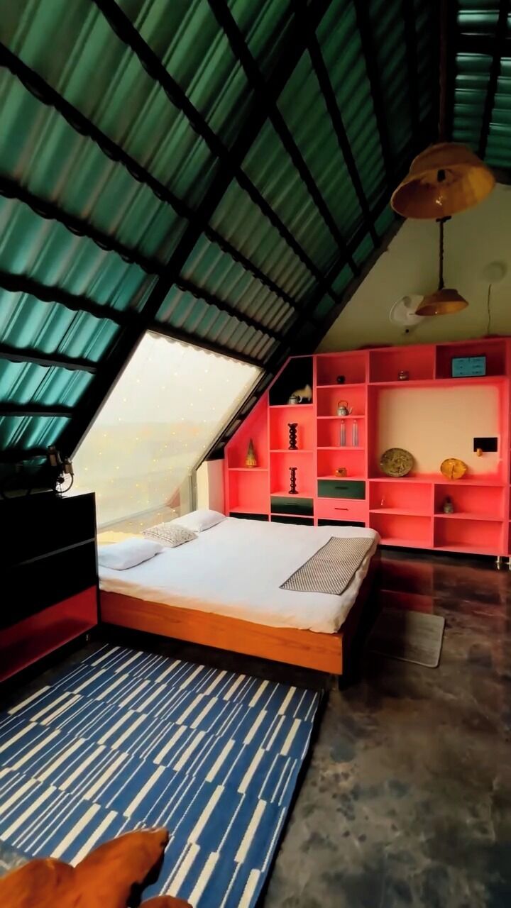 Un dormitorio con techo inclinado, una cama, un tapete azul con estampado y un estante rojo con diversos objetos.