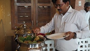 A person serving food from a stainless steel pot with a lid.