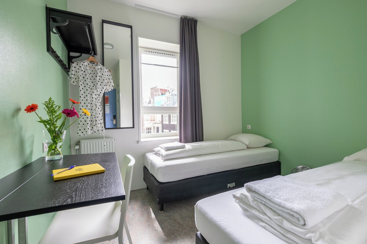 A hotel room with a bed, a desk with a chair, a mirror, and a window with curtains.