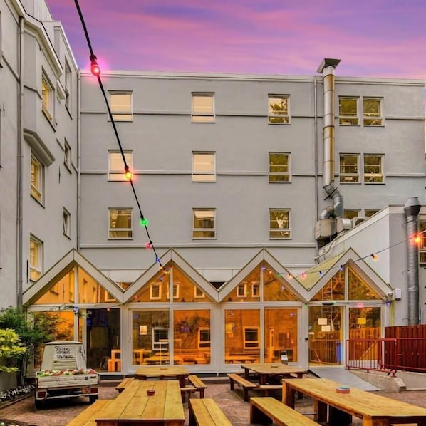 A building with a flat facade, multiple windows, and a series of wooden tables and benches in the foreground. - Hans Brinker Hostel Amsterdam (Amsterdam)