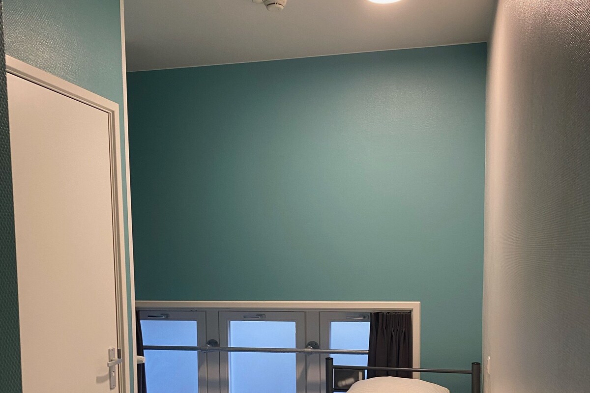 A single bed with a metal frame in a room with teal walls, a window with frosted glass, and a door.