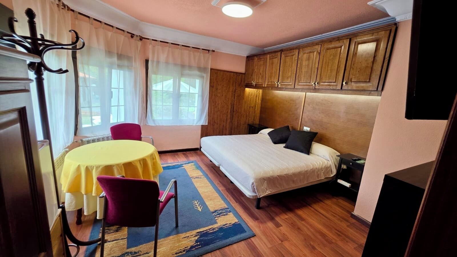 Quarto com cama, mesa de cabeceira, cadeira, mesa com toalha amarela e janela com cortinas.