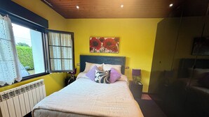 Un dormitorio con cama, ventana con cortinas, radiador, mesita de noche con lámpara y un cuadro en la pared.