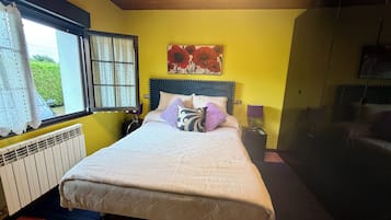 Un dormitorio con cama, ventana con cortinas, radiador, mesita de noche con lámpara y un cuadro en la pared.