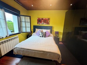 Un dormitorio con cama, ventana con cortinas, radiador, mesita de noche con lámpara y un cuadro en la pared.