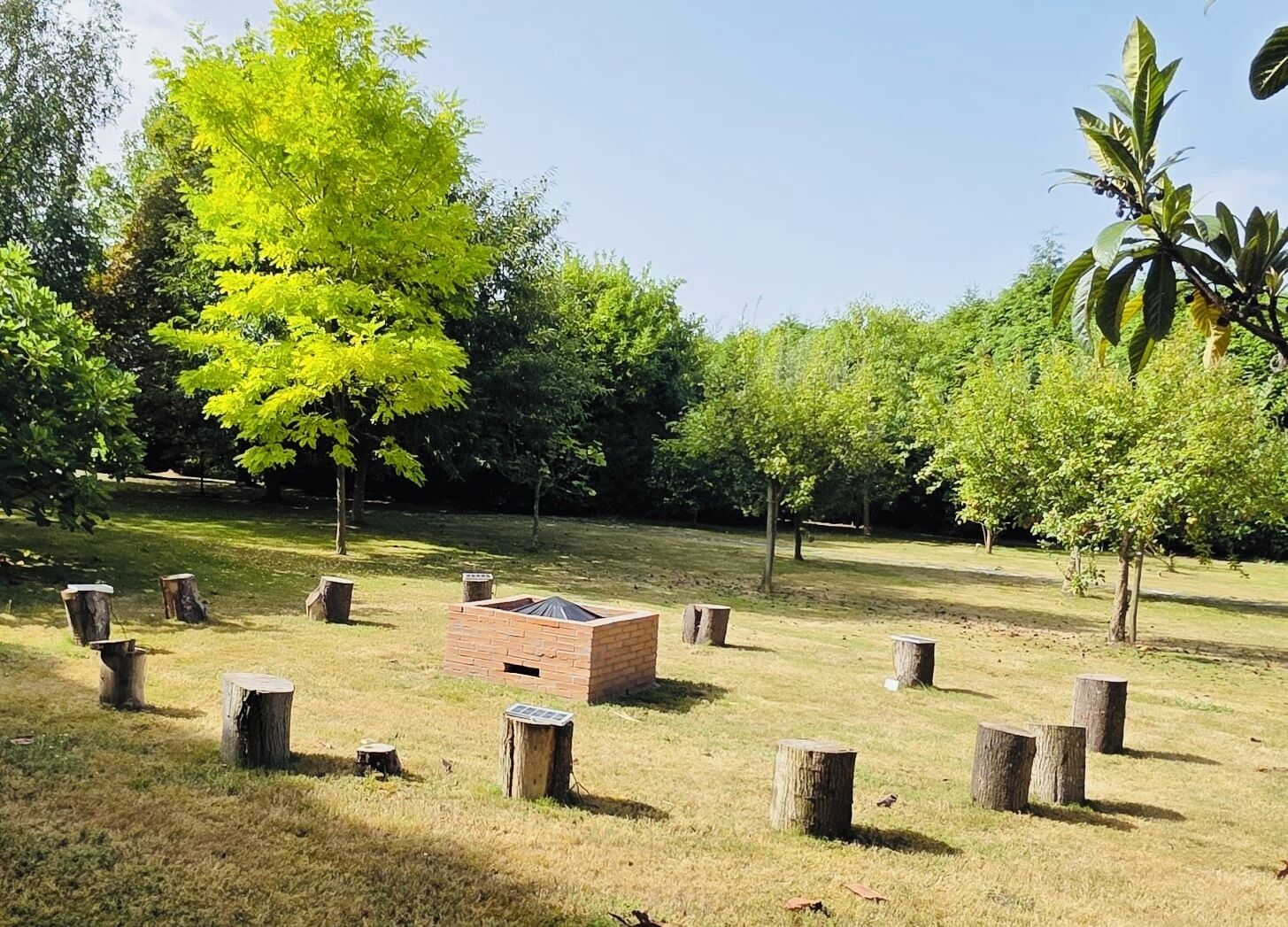 Zona verde con una estructura de ladrillo, posiblemente un área de barbacoa o para picnic, rodeada de árboles y troncos de madera.
