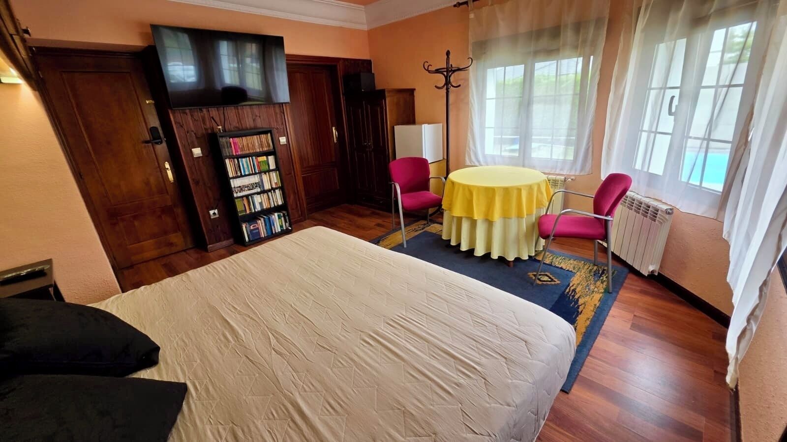 A bedroom with a bed, a bookshelf, a round table with chairs, and a window with curtains.