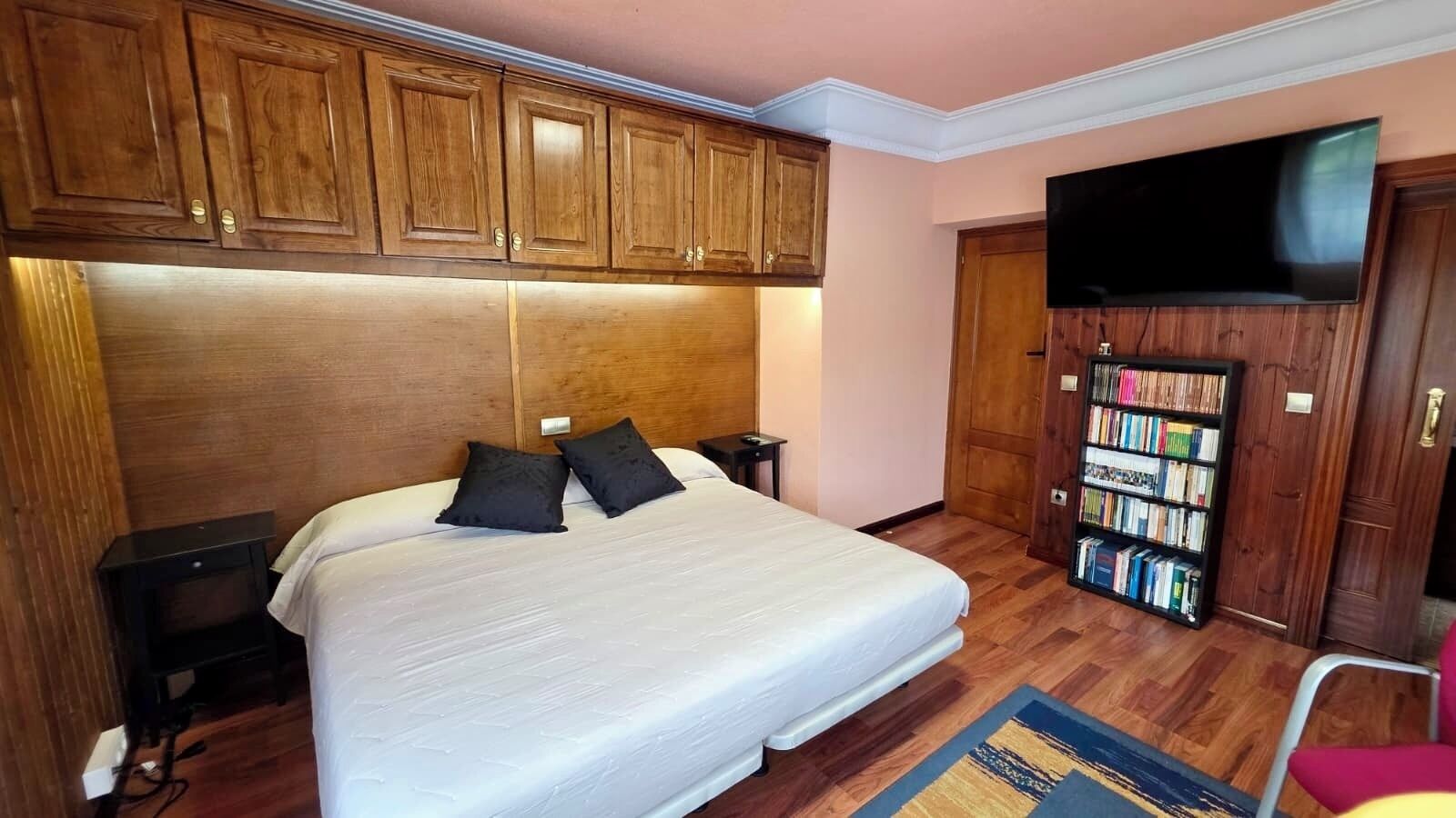 Habitación de hotel con cama, cabecero de madera, mesitas de noche, estantería y televisión.