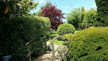 Un sentier en bois traverse un jardin bien entretenu, où l’on trouve divers arbustes et un grand arbre aux feuilles rouges.
