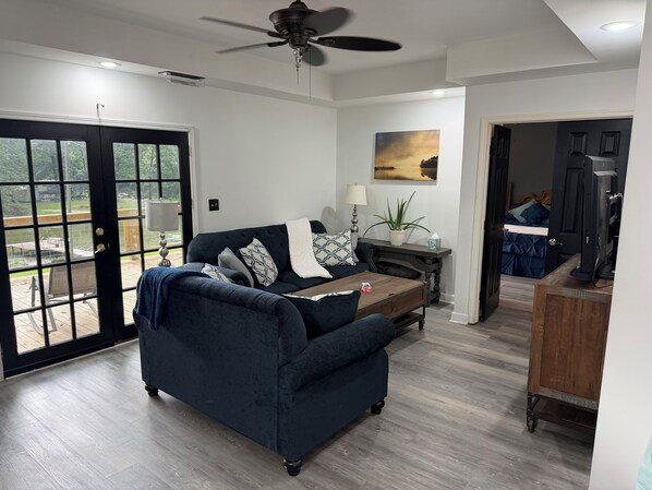 A living room with a dark blue sofa, a wooden coffee table, a ceiling fan, and a view through a glass door to an outdoor deck.