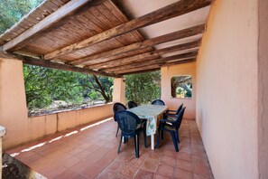 An outdoor patio with a wooden ceiling, a table with chairs, and a view of greenery.