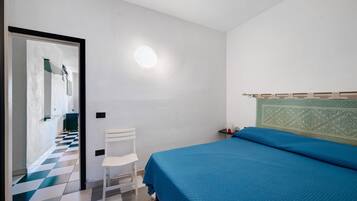 Une chambre avec un sol à carreaux, un lit recouvert d’une couverture bleue, une chaise blanche et un luminaire fixé au mur.