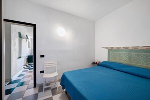 A bedroom with a checkered floor, a bed with a blue cover, a white chair, and a wall-mounted light fixture.