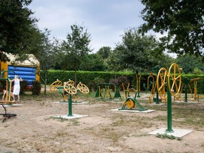 Ein Außenbereich zum Training mit verschiedenen Fitnessgeräten, darunter Spinning-Räder und Kraftgeräte, umgeben von Bäumen und einer Rasenfläche.