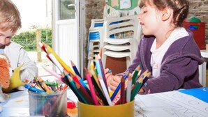 Two children at a table with various colored pencils and crayons. There are plastic chairs stacked in the background, and a window with a view of greenery outside. - Mobile home with AC - 6 guests (Sainte-Reine-de-Bretagne)