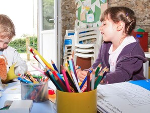 Zwei Kinder sitzen an einem Tisch mit verschiedenen Buntstiften und Wachsmalstiften. Im Hintergrund sind Plastikstühle gestapelt und durch ein Fenster ist Grünanlage zu sehen.