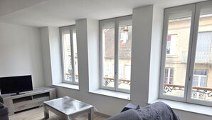 Una sala de estar con un televisor de pantalla plana, una mesa de centro de madera y un sofá. Hay ventanas amplias con vistas a edificios.