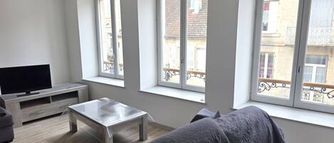 Una sala de estar con un televisor de pantalla plana, una mesa de centro de madera y un sofá. Hay ventanas amplias con vistas a edificios.