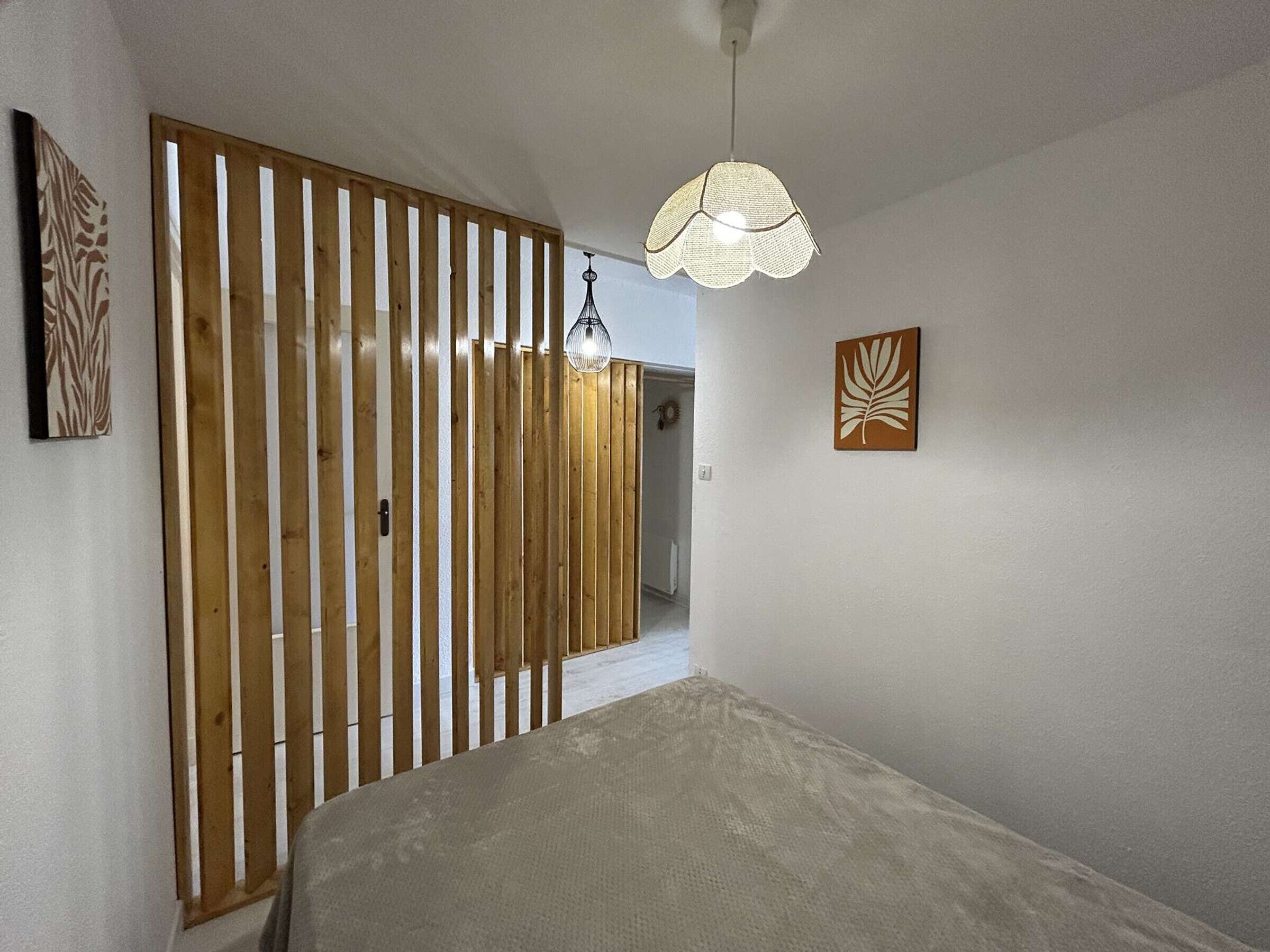 Une chambre comprenant un lit, une cloison en lattes de bois, un luminaire suspendu et deux œuvres d’art fixées au mur.