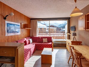 A cozy room with a wooden interior, a red patterned sofa, a wooden dining table, and a view of a snowy mountain landscape.