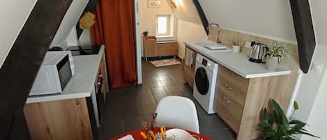 Un coin repas avec une table rouge dressée pour six personnes, une cuisine équipée d’un lave-linge et un espace en mezzanine avec des poutres apparentes.