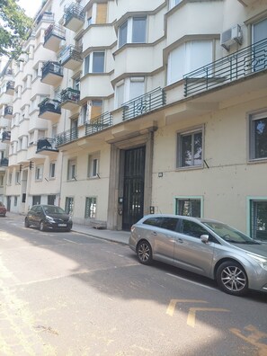 Un edificio residencial de varios pisos con balcones, una entrada con portón y coches estacionados en la calle.