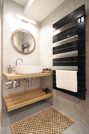 Un bagno moderno con un lavandino bianco, uno specchio rotondo, un porta asciugamani nero e una mensola in legno.