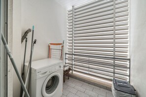 Ein Waschraum mit einer Waschmaschine, einem Wäscheständer und einem Fenster mit Jalousien.