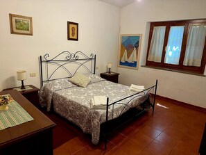 A bedroom with a metal bed frame, a bedside table with a lamp, a window with curtains, and artwork on the walls.
