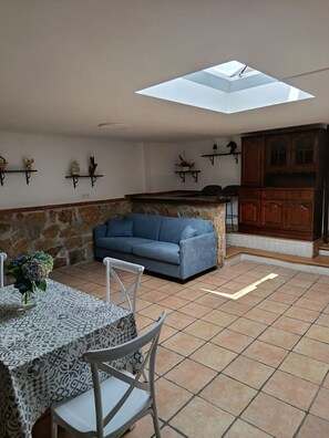 A living area with a stone wall, a blue sofa, a dining table with chairs, and a kitchen area with wooden cabinets. - Atalaya 4001 Ground-Floor Apartment (Arnuero)