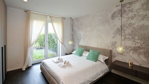 Quarto moderno com uma cama grande, duas almofadas, uma mesa de cabeceira, uma luminária na parede e vista para o verde pela janela.